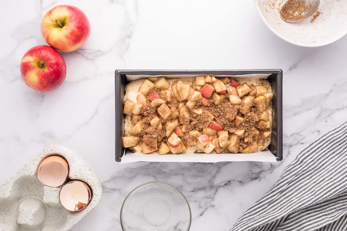 An unbaked apple fritter bread topped with chopped apples and spices in a loaf pan on a white background with apples and other extra ingredients around it.