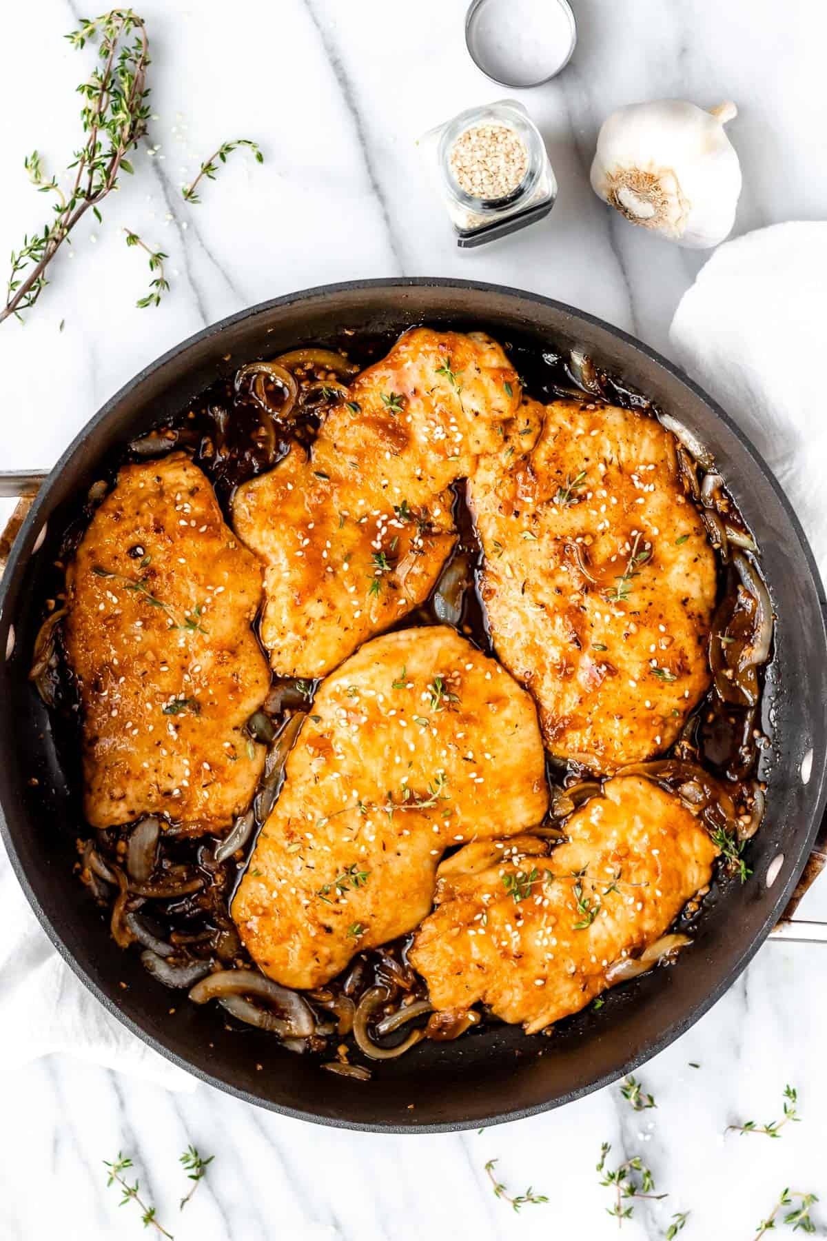 Five cooked chicken breasts in apricot sauce with onions in a black skillet with fresh thyme, garlic and a container of sesame seeds around it.
