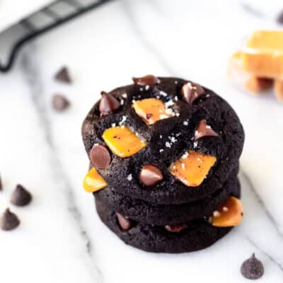 A stack of 3 dark chocolate chip salted caramel cookies on a marble background with chocolate chips and caramels around them.