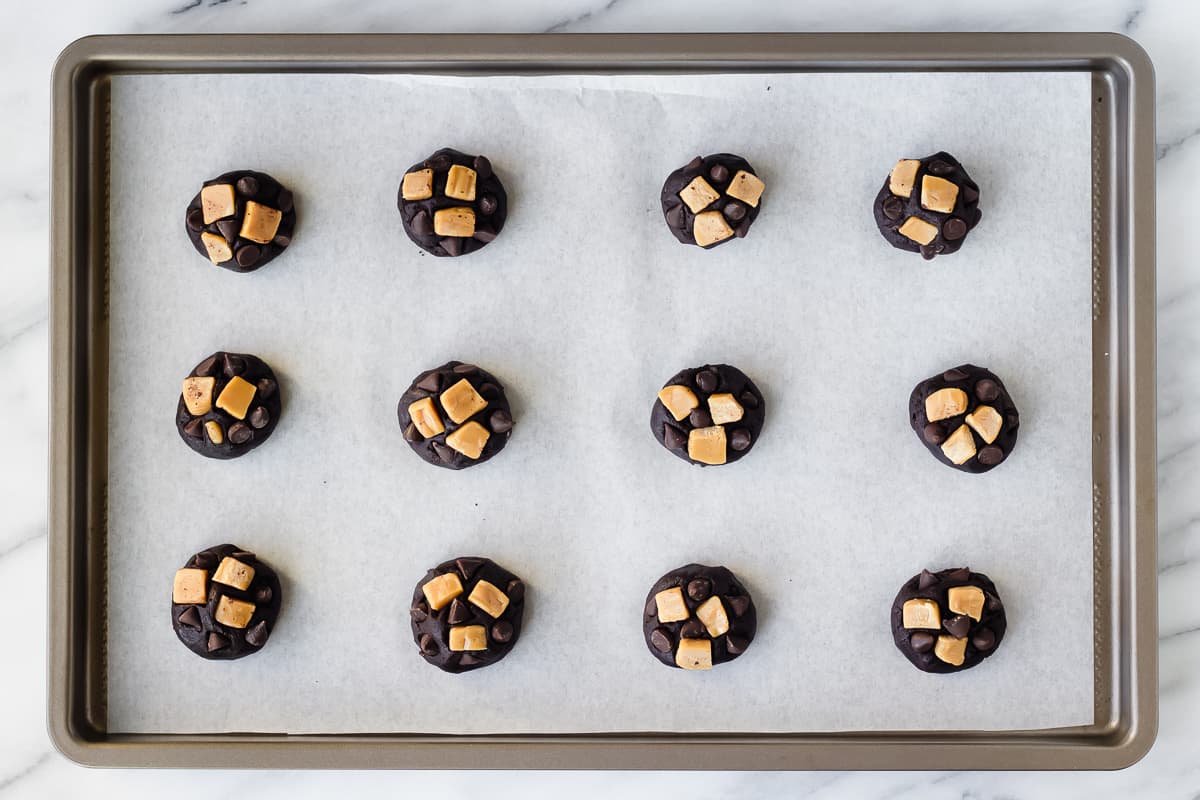 12 dark chocolate dough balls on a parchment paper lined baking sheet with caramel pieces and chocolate chips pressed onto the tops of each one.