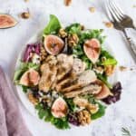 A salad with figs, walnuts and feta cheese on a white plate with extra ingredients around it.