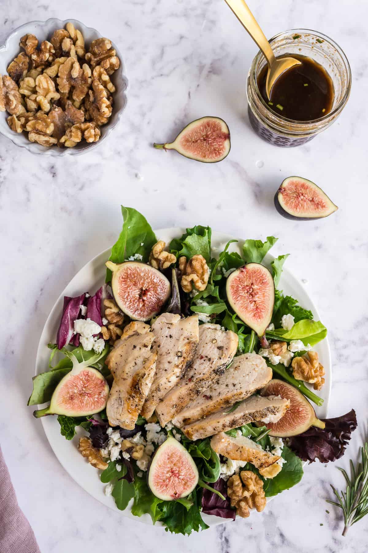 A salad with figs, walnuts and feta cheese topped with chicken on a white plate with extra ingredients around it.