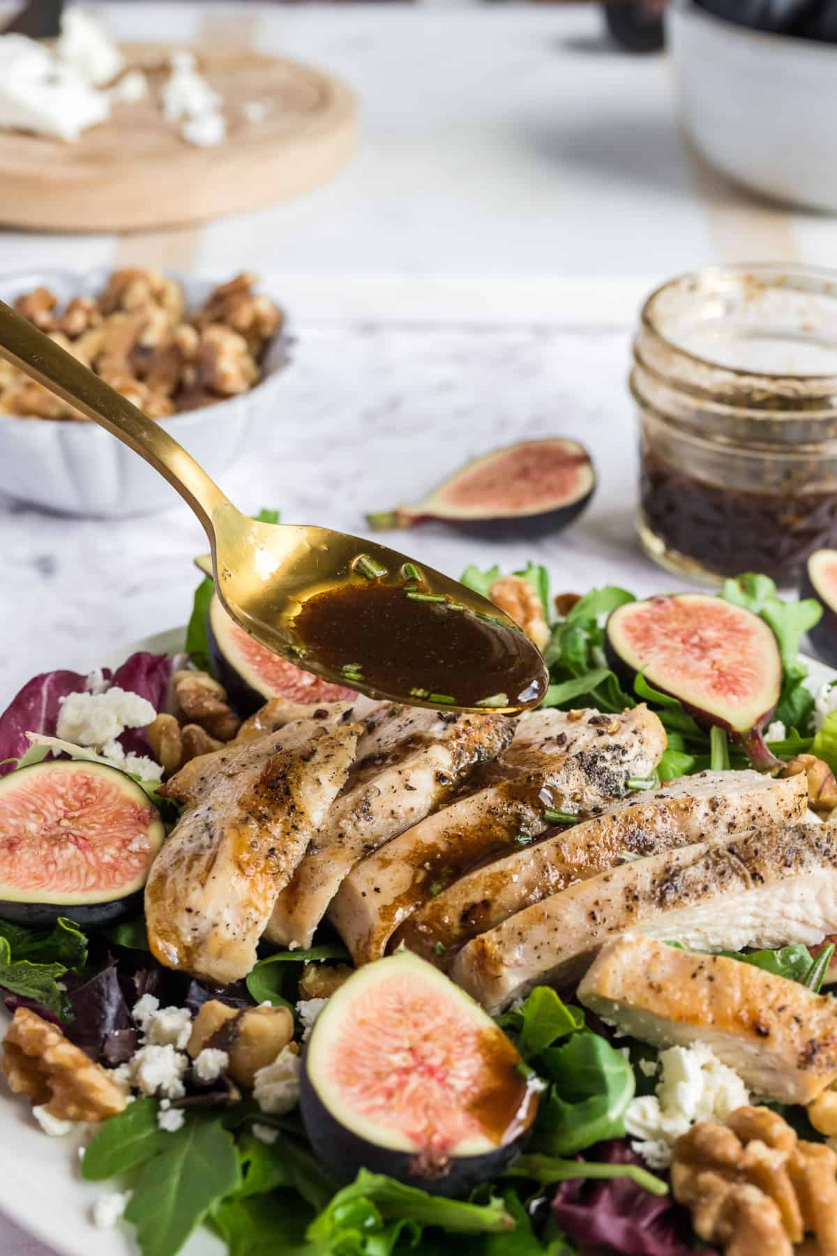 Balsamic dressing being drizzled over a fig and chicken salad with a spoon with a jar of dressing and bowl of walnuts in the background.