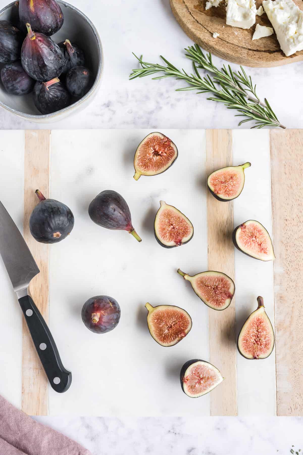 Whole and halved figs on a cutting board with a knife with a bowl of figs and rosemary in the background.