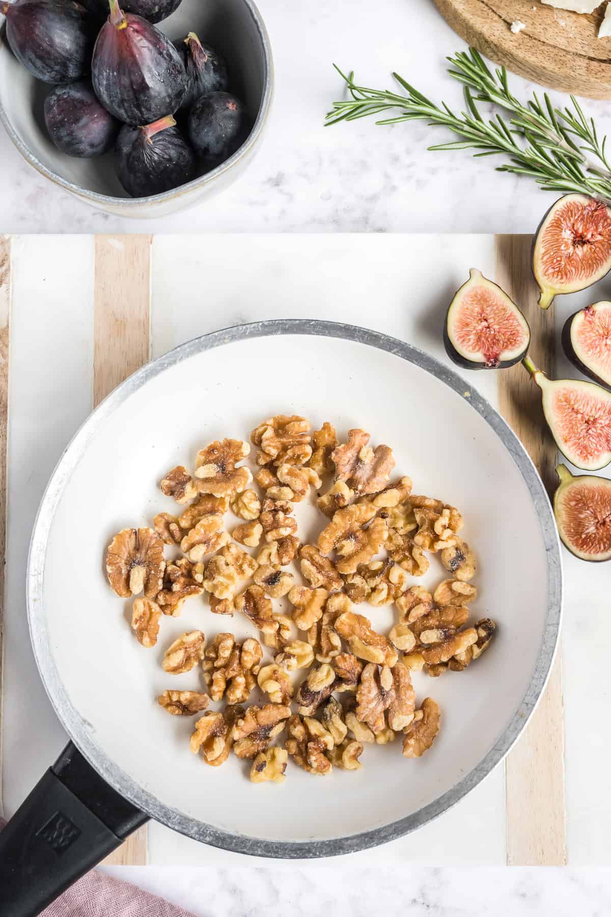 Toasted walnuts in a white skillet with whole and halved figs and rosemary around it.