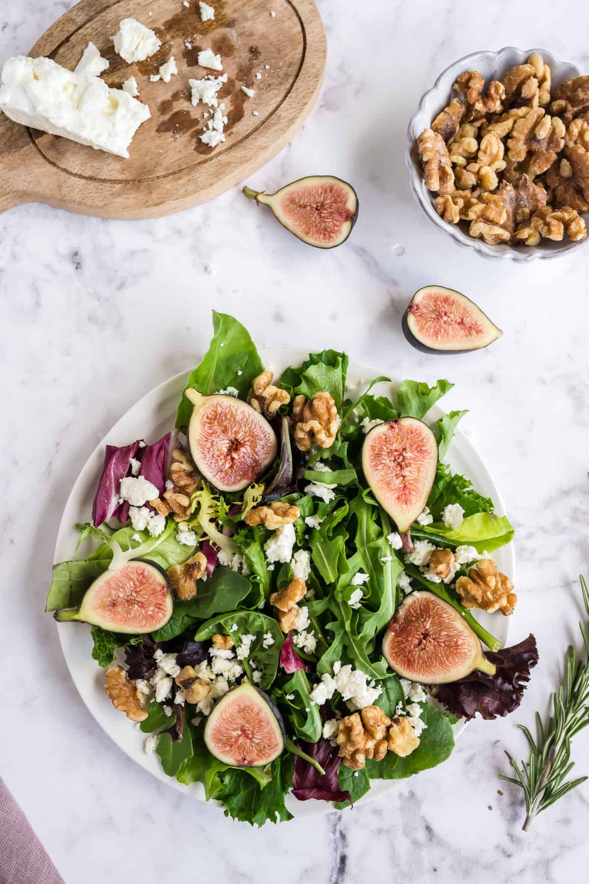 A salad with figs, walnuts and feta cheese on a white plate with extra ingredients around it.