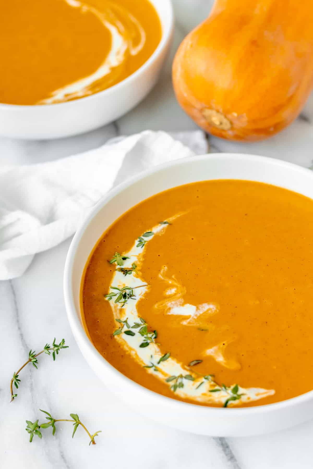 Close up of a bowl of honeynut squash soup topped with cream and thyme with a second bowl and a honeynut squash partially showing in the background.