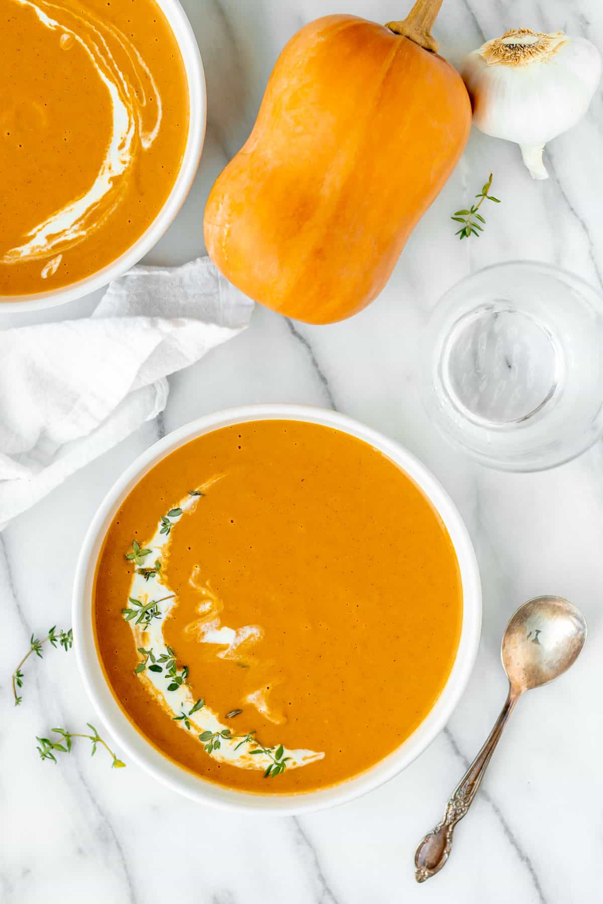 Overhead of a bowl of honeynut squash soup with a second bowl partially showing, a honeynut squash, garlic head and spoon around it.