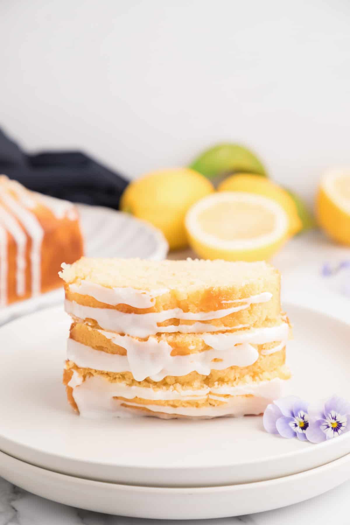 Three sliced of lemon drizzle cake stacked on top of each other on a white plate with lemons and a towel in the background.