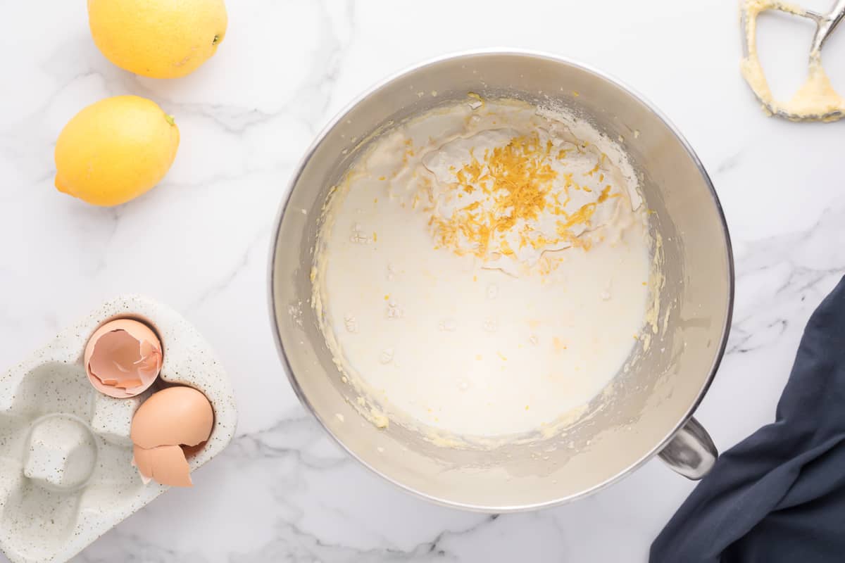 Lemon cake batter with some flower and lemon zest on the top in a mixing bowl with lemons, egg shells and a towel in the background.