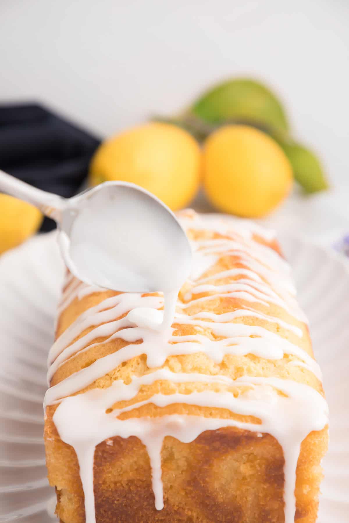 Glaze being drizzled over the top of a lemon cake with lemons and a towel in the background.