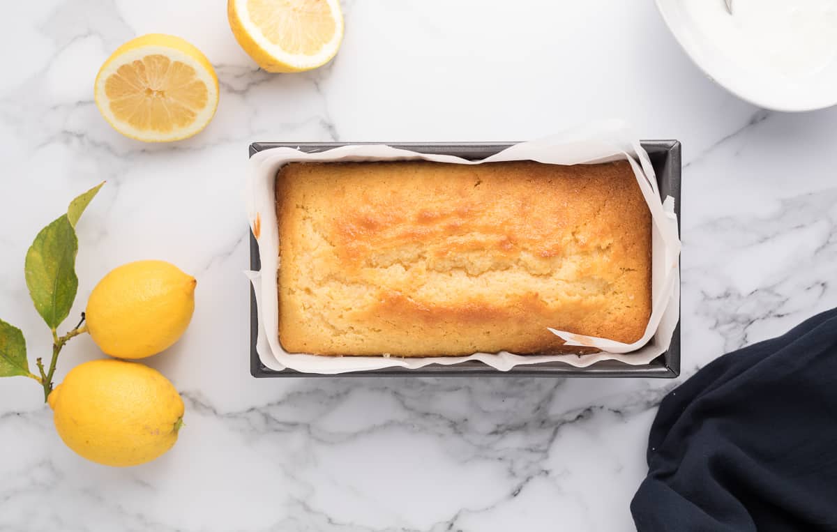 A baked lemon cake in a parchment paper lined loaf pan with lemons and a towel in the background.