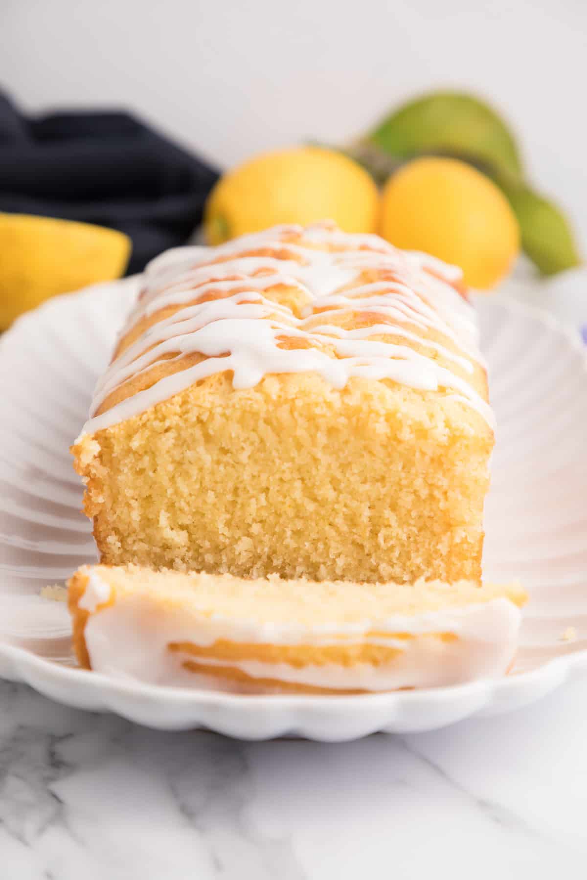 A lemon drizzle cake facing the front with one slice cut leaning forward on a white plate with lemons and a towel in the background.