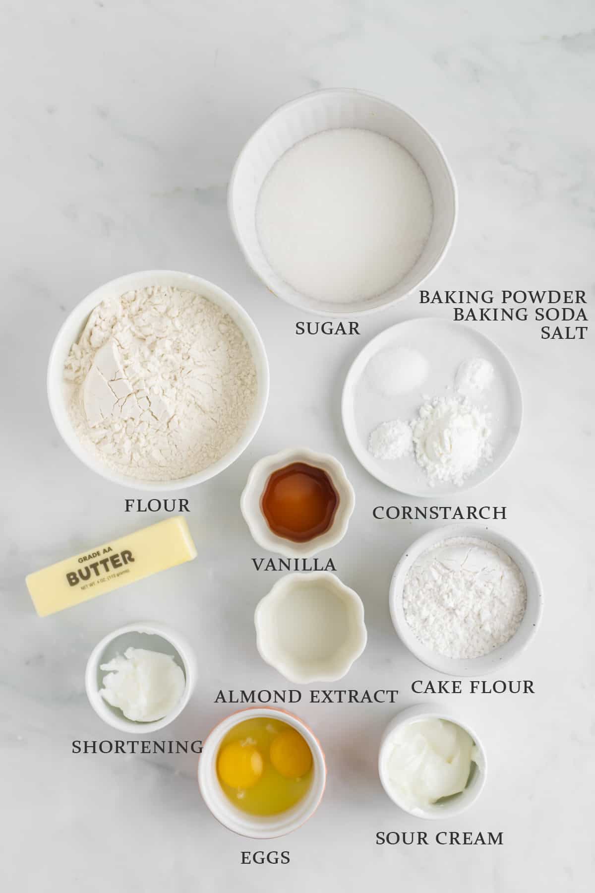 Ingredients to make sugar cookies in small bowls on a white background. Text overlay reads the ingredient names.