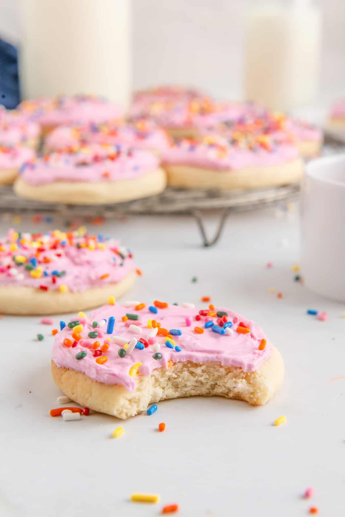 A Lofthouse sugar cookie with pink frosting and rainbow sprinkles that has a bite taken out of it with more cookies in the background.