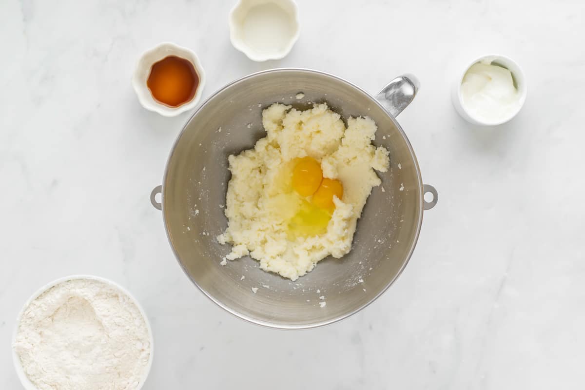 Butter and sugar creamed together in a mixing bowl with eggs on top and other ingredients in small bowls around it.