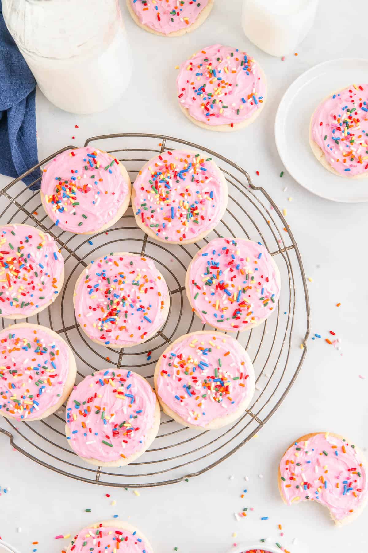 Overhead of sugar cookies with pink frosting and rainbow sprinkles on a cooling rack with more cookies and sprinkles around it.