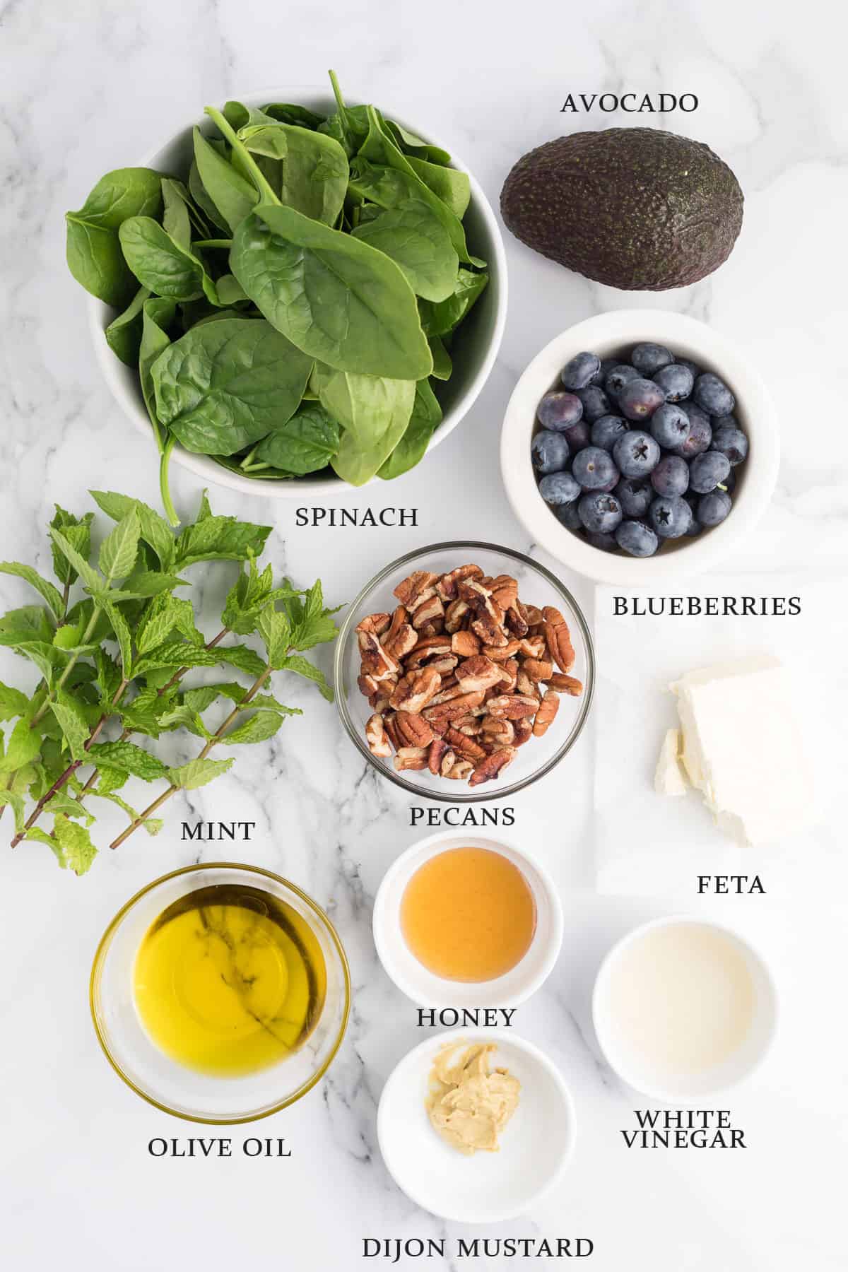Ingredients needed to make a spinach blueberry salad in bowls or on a white surface with text overlay.