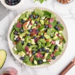 Overhead of a spinach blueberry salad with pecan, blueberries, avocado, feta, mint and blueberry dressing on a white plate with extra ingredients around it.