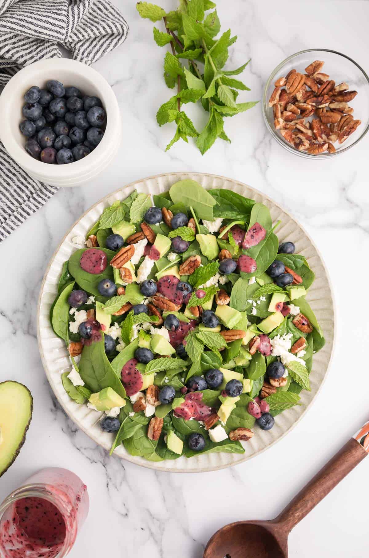 Overhead of a spinach blueberry salad with pecan, blueberries, avocado, feta, mint and blueberry dressing on a white plate with extra ingredients around it.