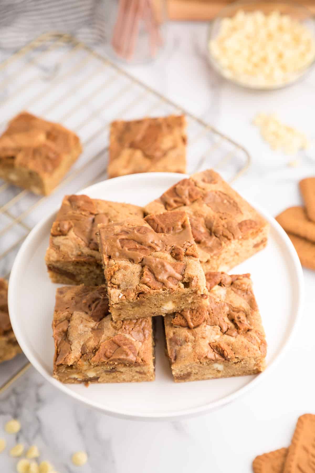 Biscoff blondies on a white plate with more on a cooling rack and a bowl of white chocolate chips in the background.