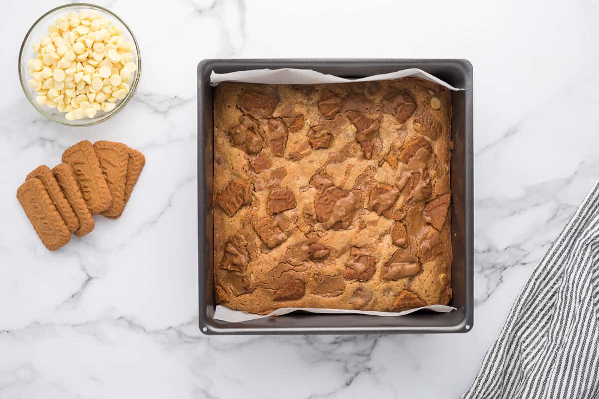 Baked Biscoff Blondies in a square baking pan with cookies and white chocolate chips in a bowl next to it.