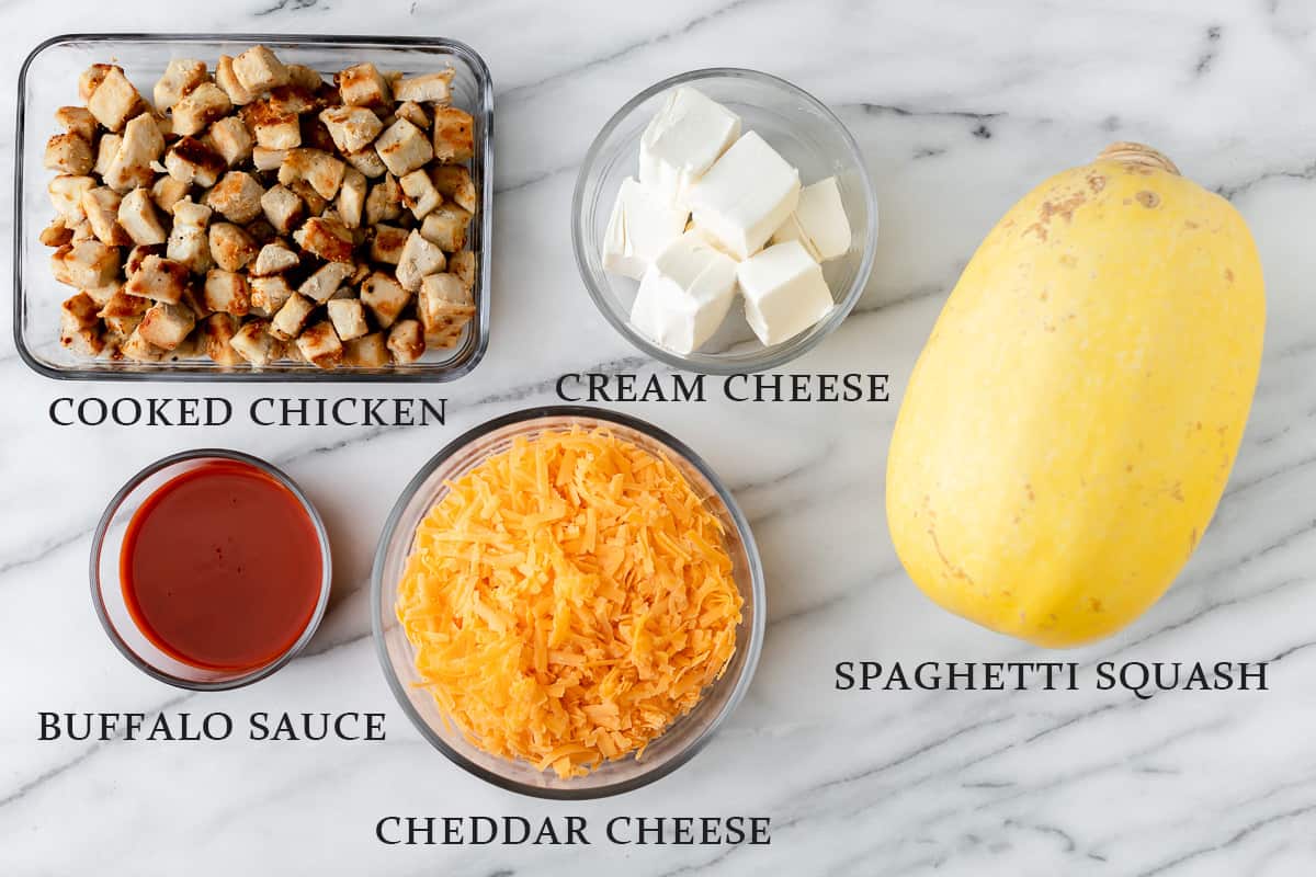 Ingredients needed to make creamy buffalo chicken spaghetti squash casserole on a marble background with text overlay.