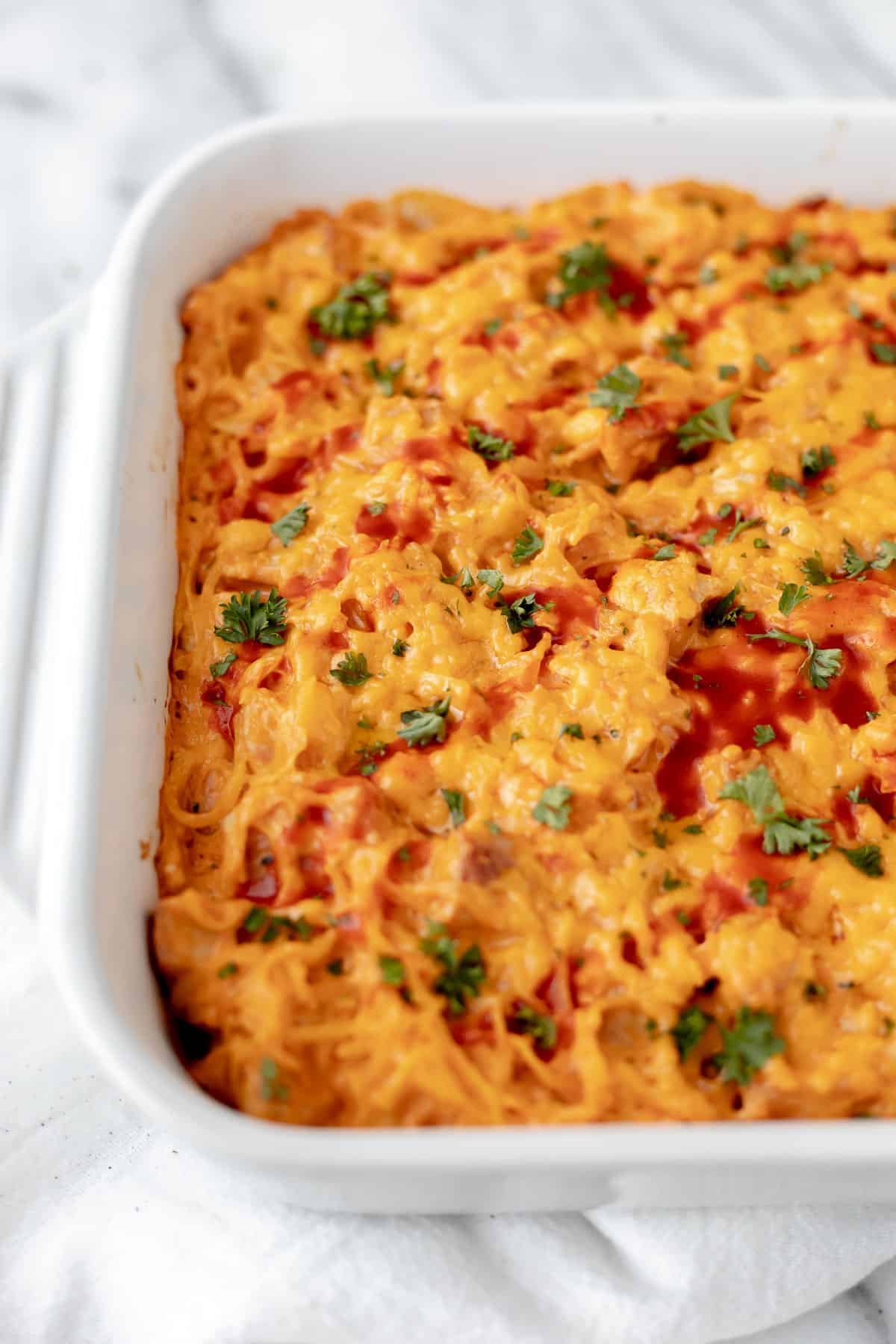 Close up of a creamy buffalo chicken spaghetti squash casserole topped with buffalo sauce and parsley in a white baking dish.