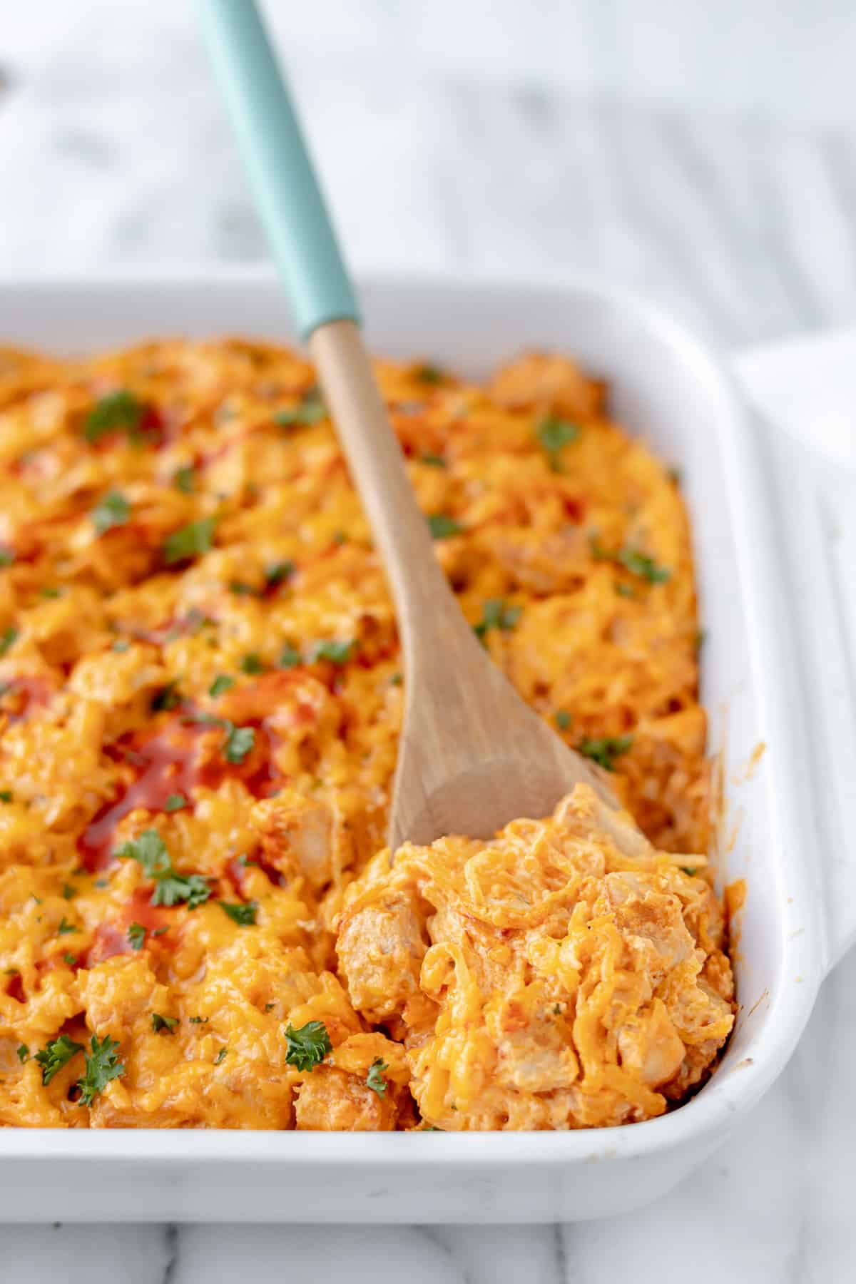 Close up of a wood spoon lifting up some buffalo chicken spaghetti squash casserole in a white baking dish.