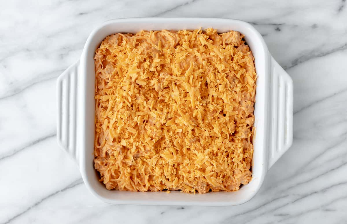 A casserole topped with shredded cheddar cheese in a white, square baking dish.