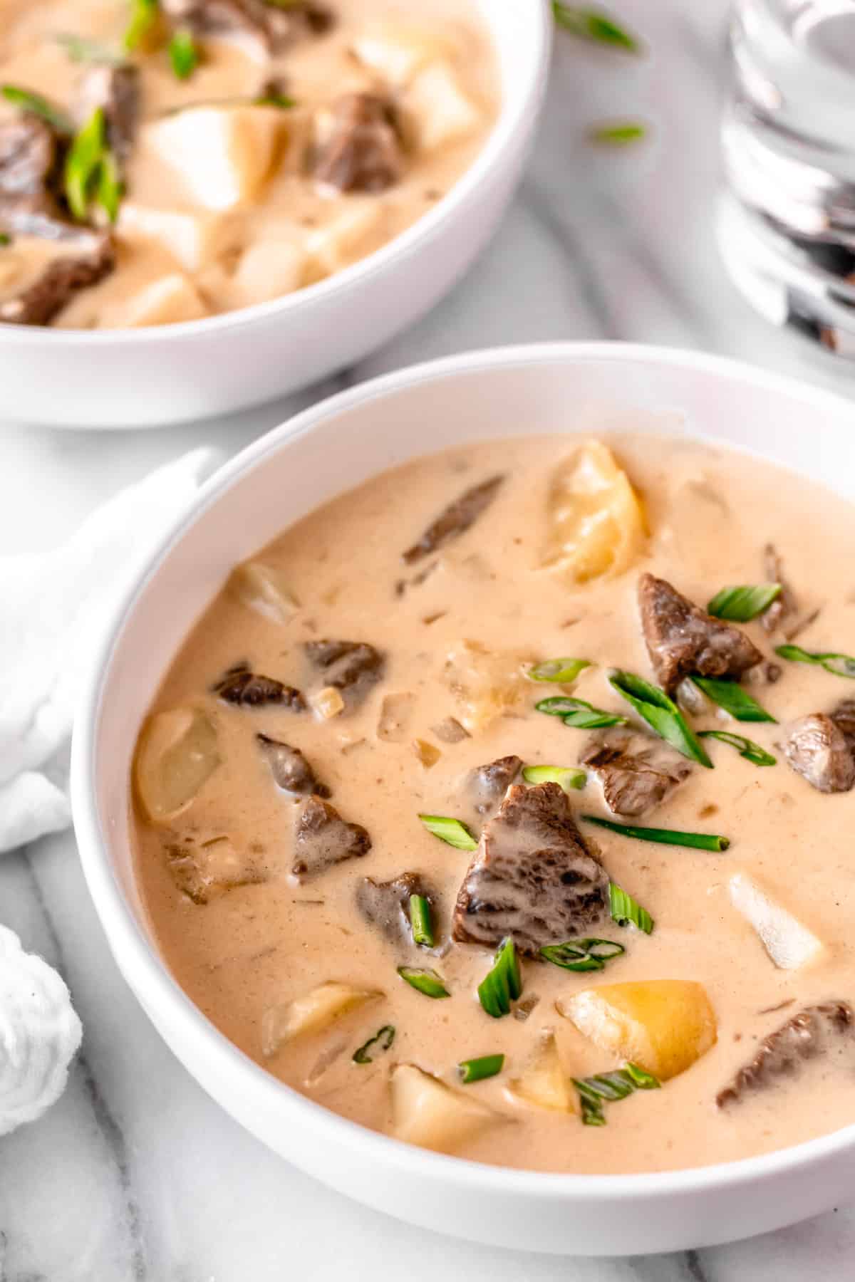 Two bowls of creamy steak and potato soup with a white towel and glass of water partially showing.