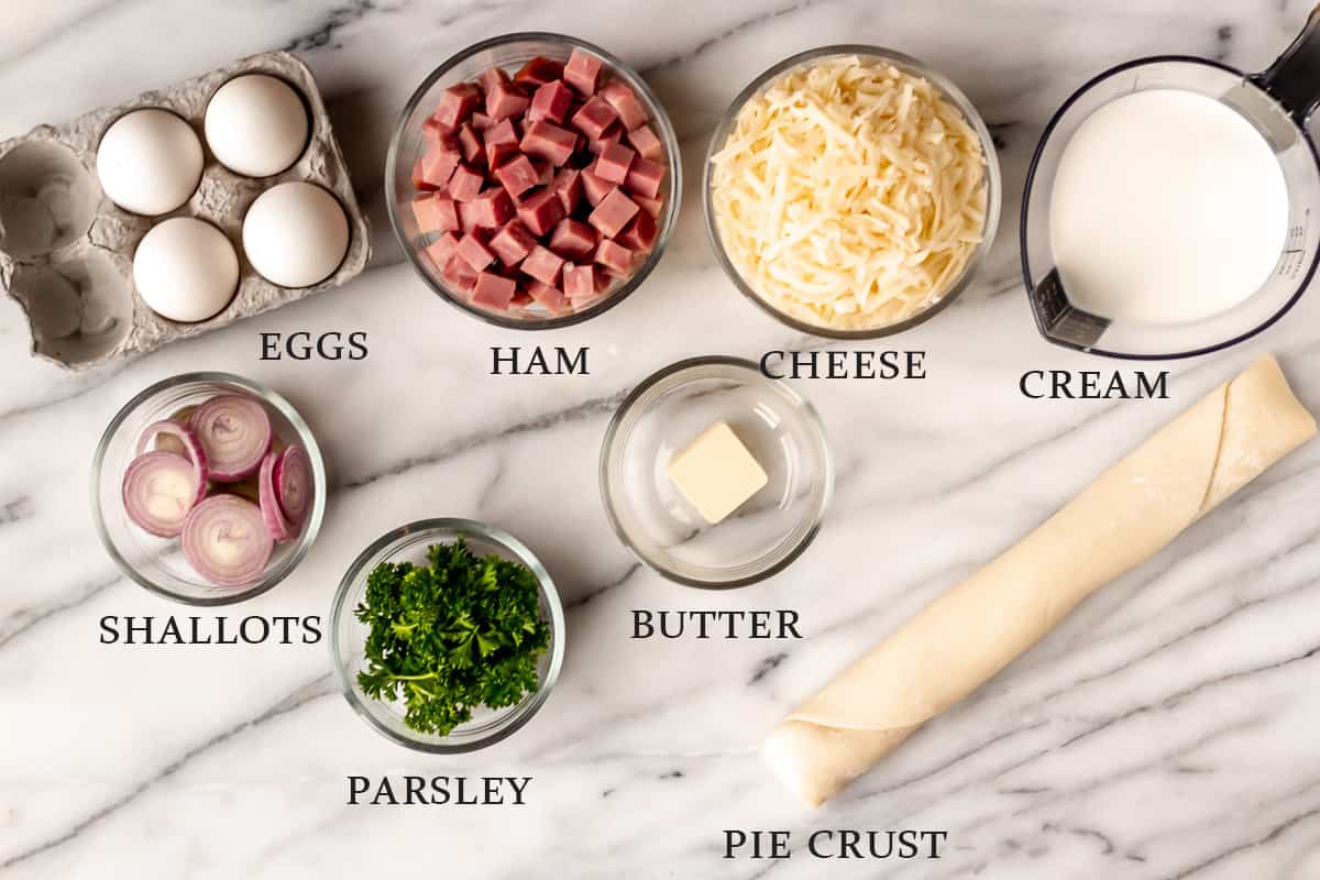 Ingredients needed to make a ham and cheese quiche on a marble background with text overlay.