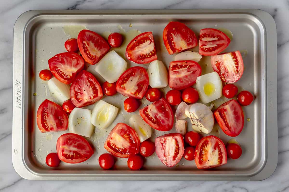 Raw chopped tomatoes, onions and a head of garlic on a baking sheet.