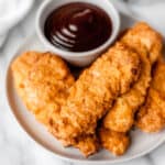 A stack of air fryer chicken tenders on a white plate with a white bowl of barbecue sauce.