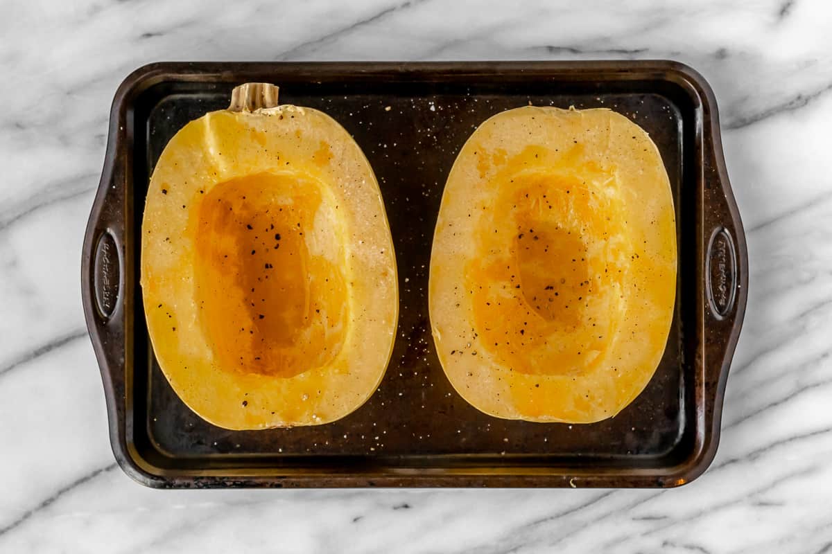 A raw spaghetti squash cut in half and seasoned with olive oil, salt and pepper on a baking sheet.