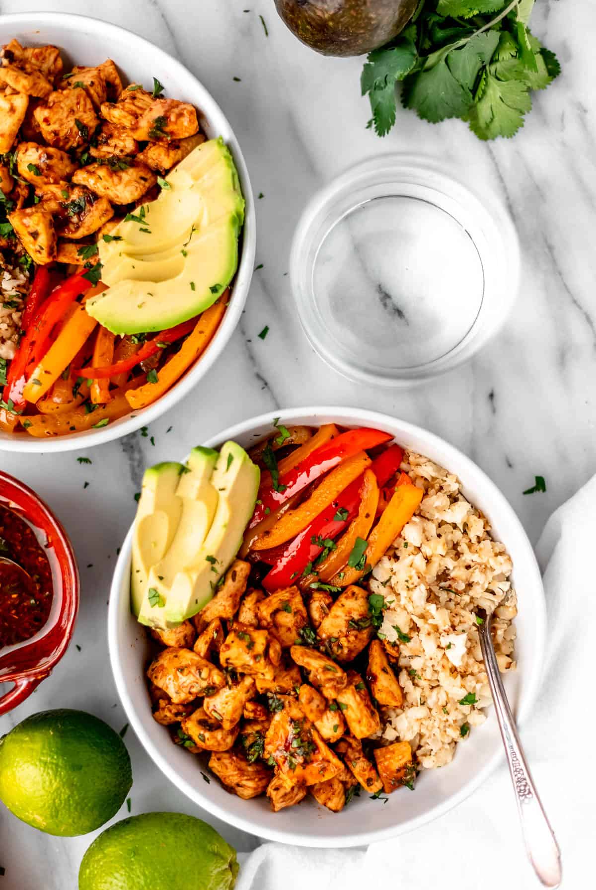 Overhead of two chicken fajita bowls filled with chicken, cauliflower rice, peppers and avocado with limes, cilantro, a glass of water and white towel around them.