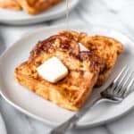 Two slices of French toast on a white plate topped with butter with syrup being drizzled on.