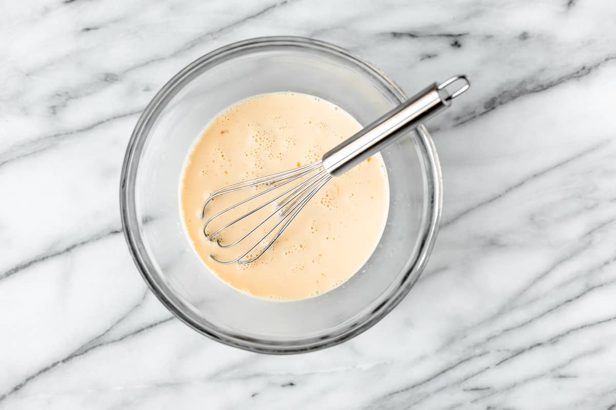 French toast custard in a clear mixing bowl with a whisk in it over a marble background.