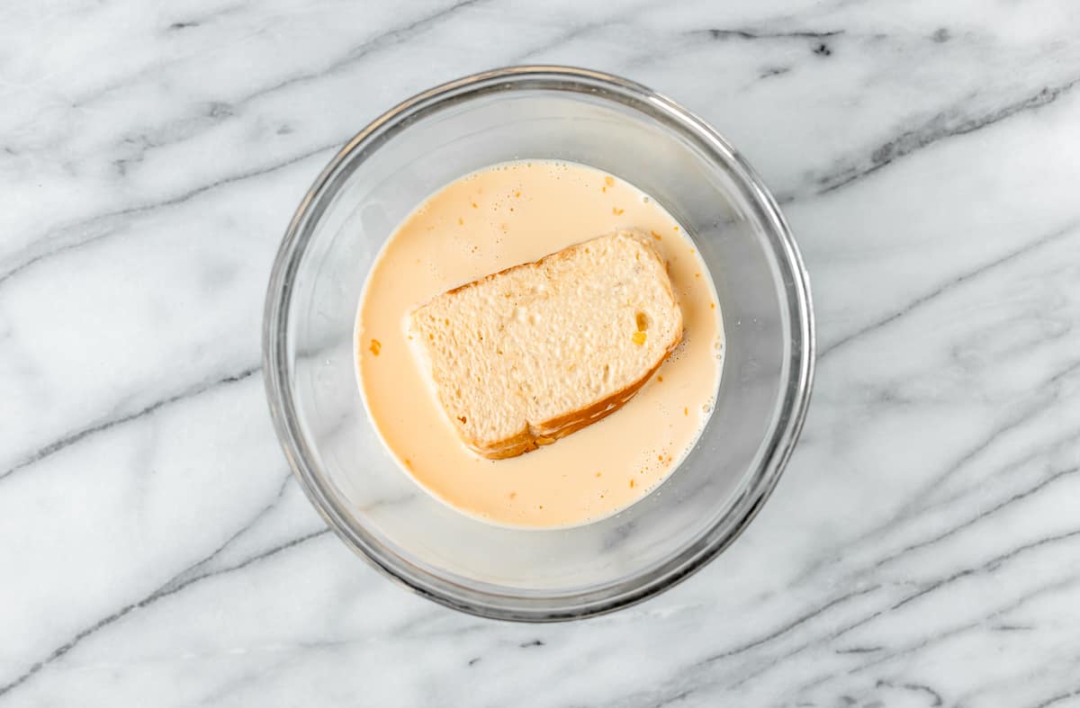 A slice of bread in egg custard in a clear mixing bowl over a marble background.