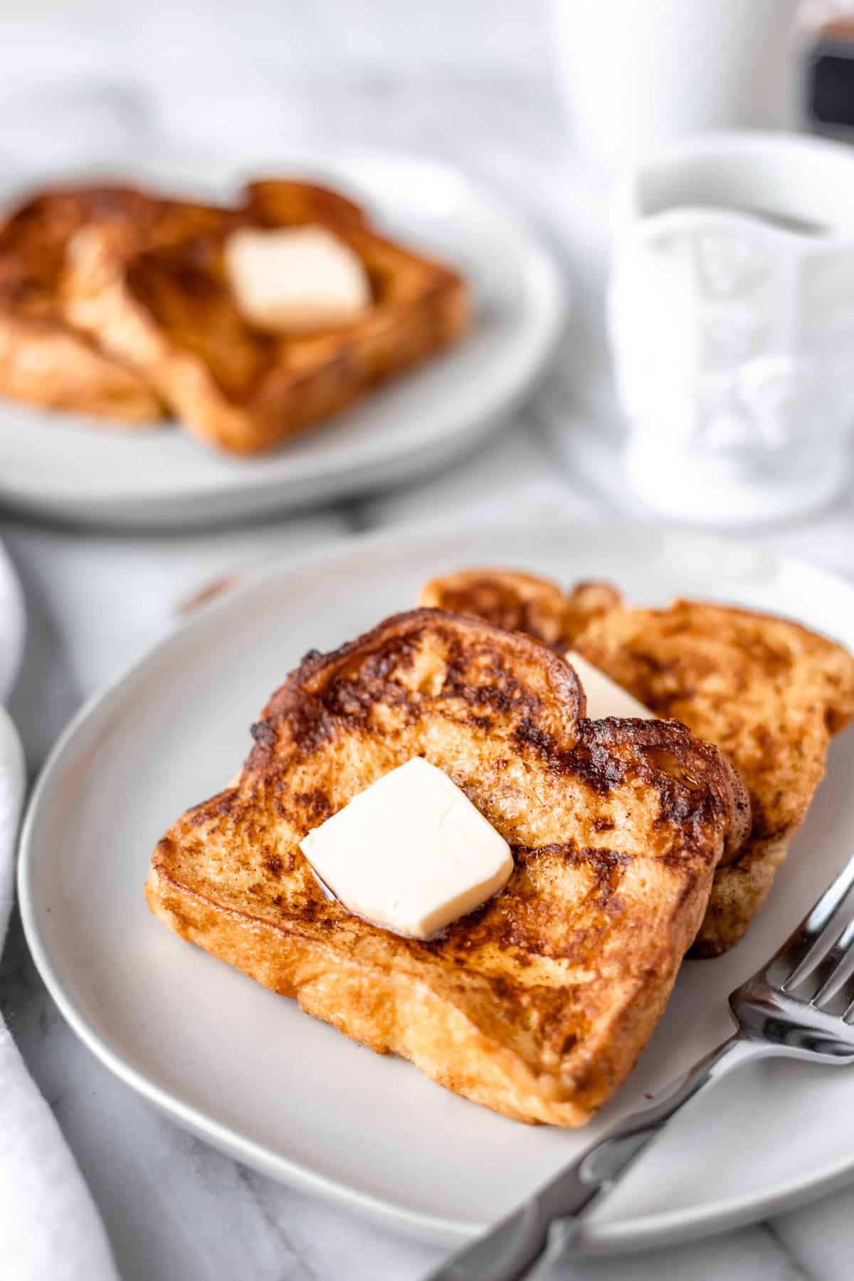 Two slices of French toast on a white plate topped with butter. With a second plate of French toast and a container of maple syrup n the background.