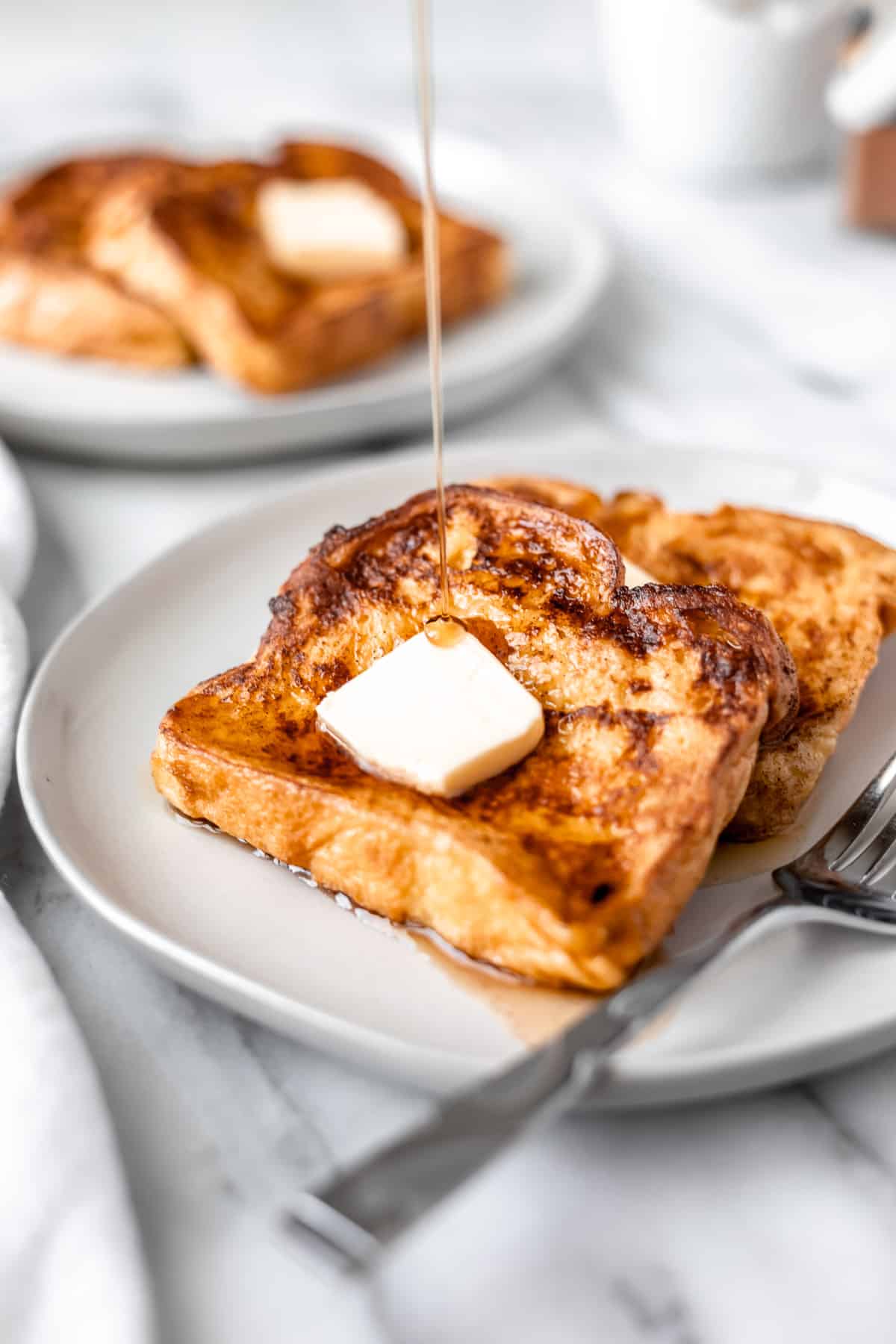 Two slices of French toast on a white plate topped with butter with syrup being drizzled on.