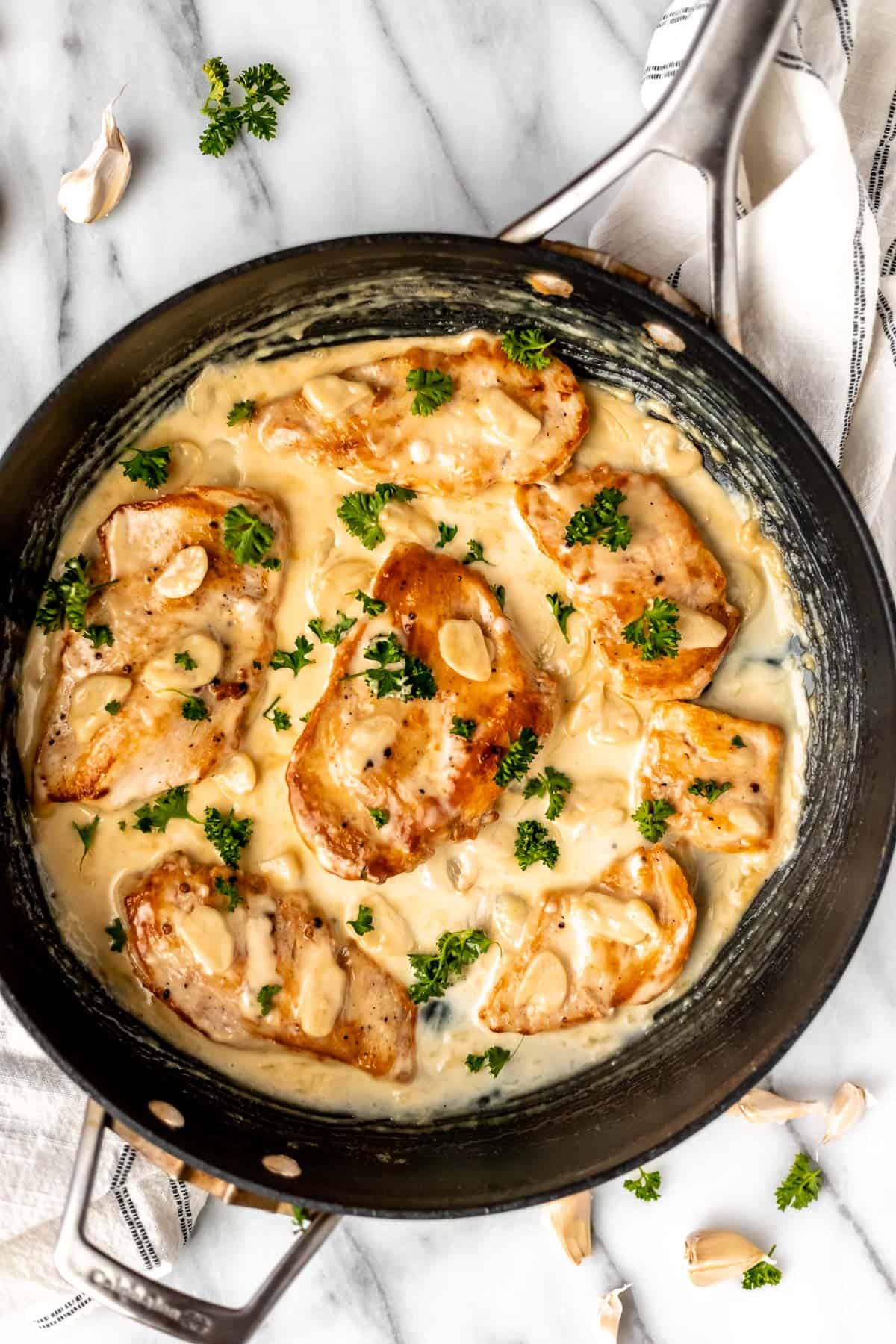 Overhead of creamy garlic chicken garnished with fresh parsley in a black skillet over a marble background.