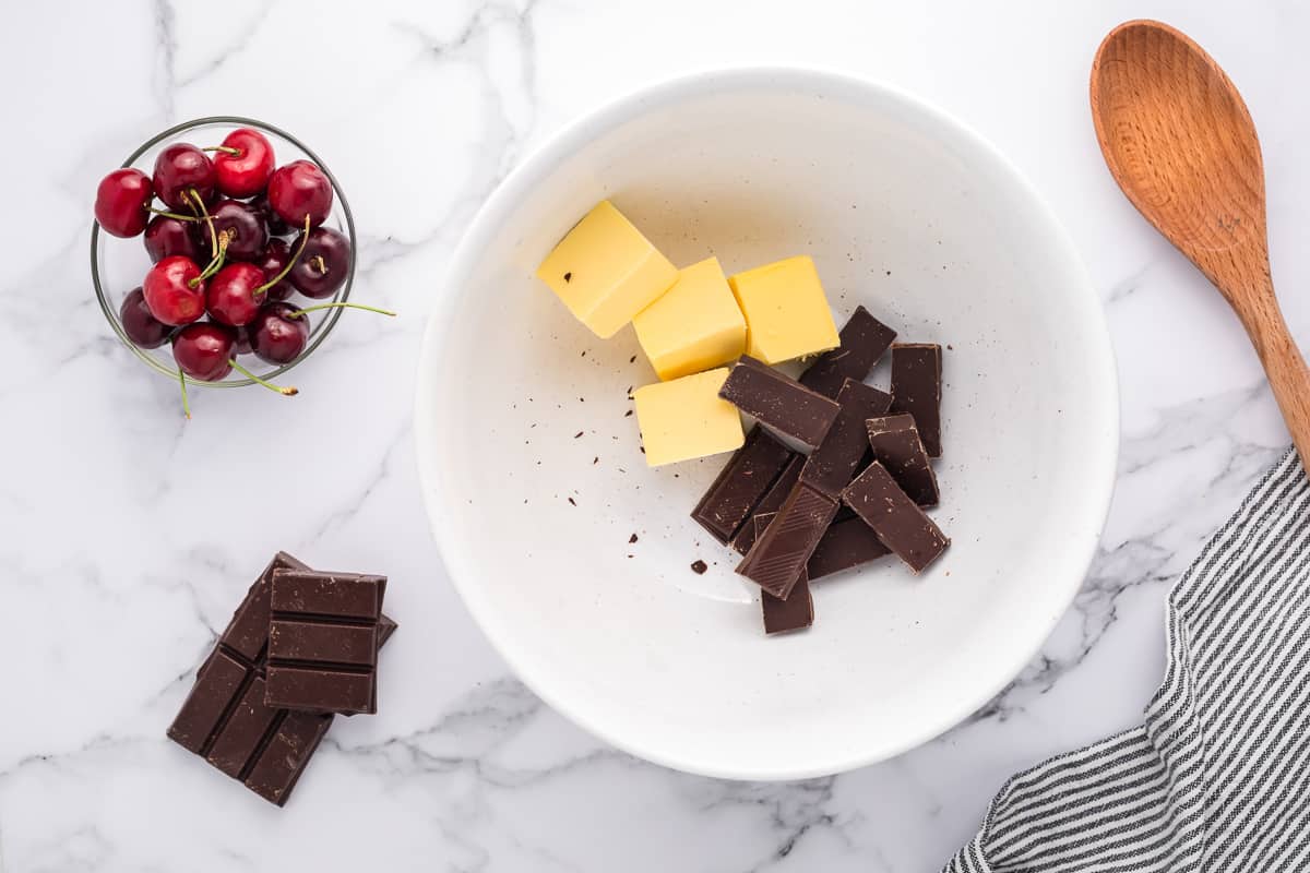 Butter and chocolate in a white bowl with a spoon, cherries, chocolate and a striped towel around it.