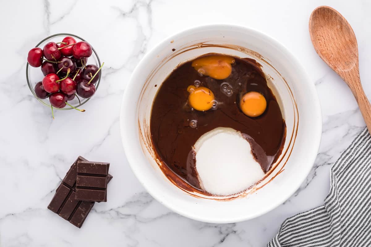 Eggs and sugar added to a chocolate batter in a white bowl with cherries, chocolate and a wood spoon around it.
