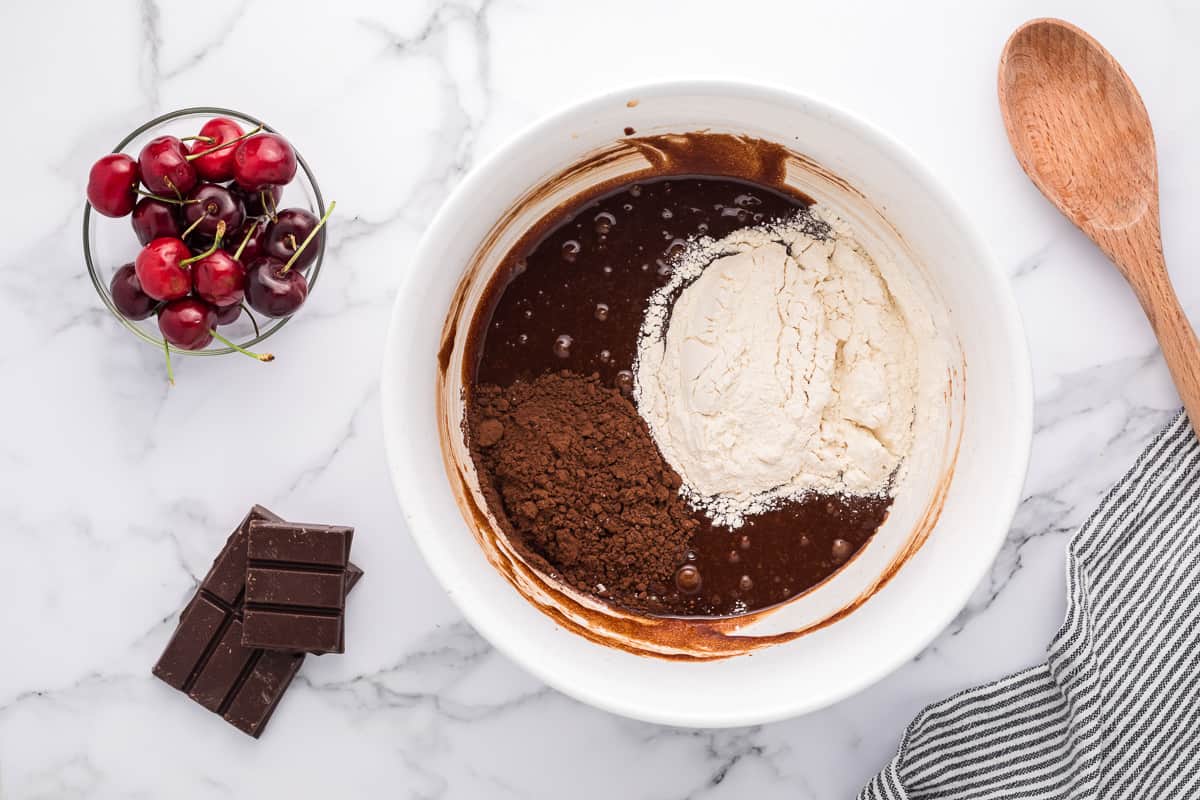 Flour and cocoa powder on top of a chocolate batter in a white bowl with cherries and chocolate around it.