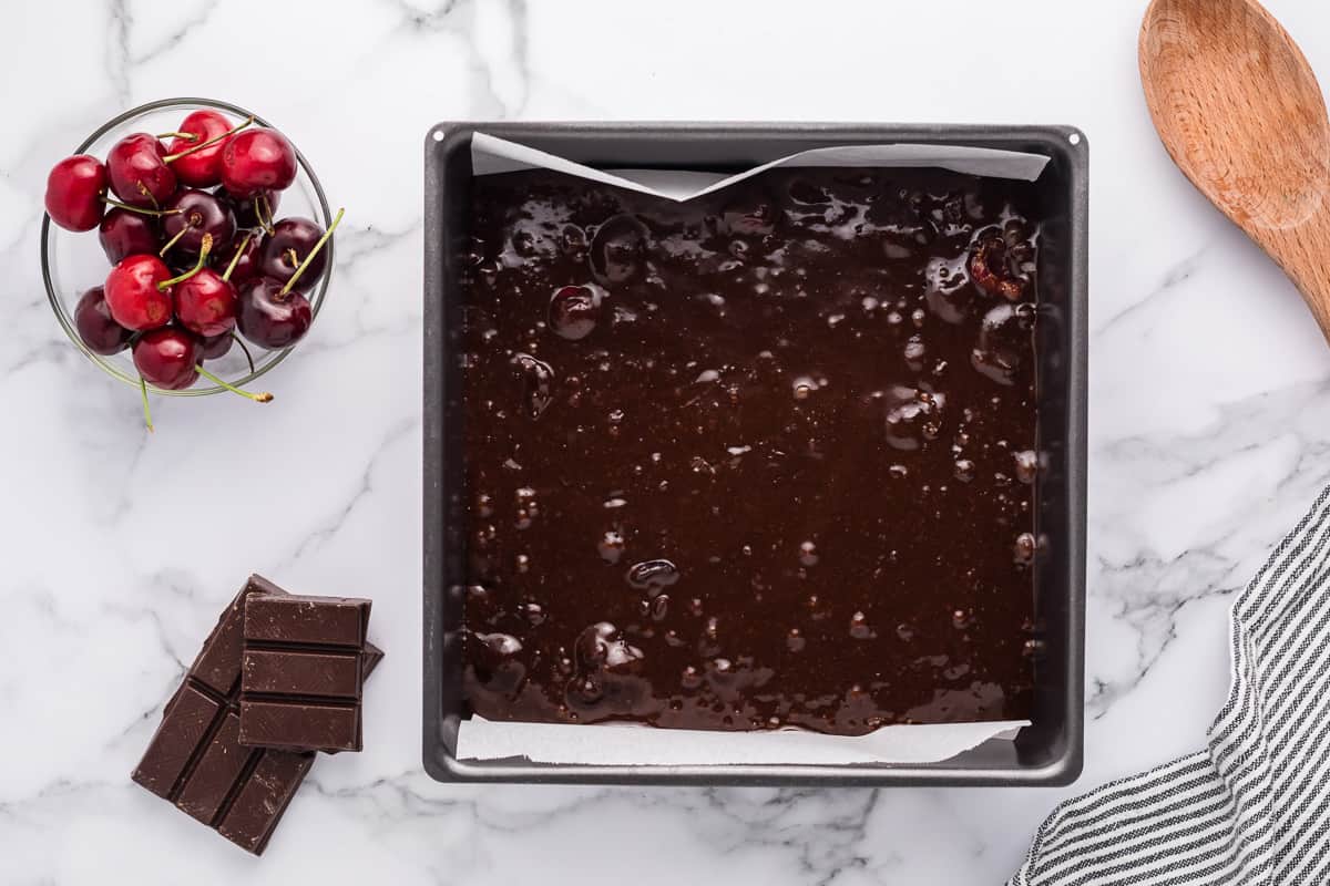 Chocolate cherry brownie batter in a square baking pan with cherries, chocolate and a wood spoon around it.