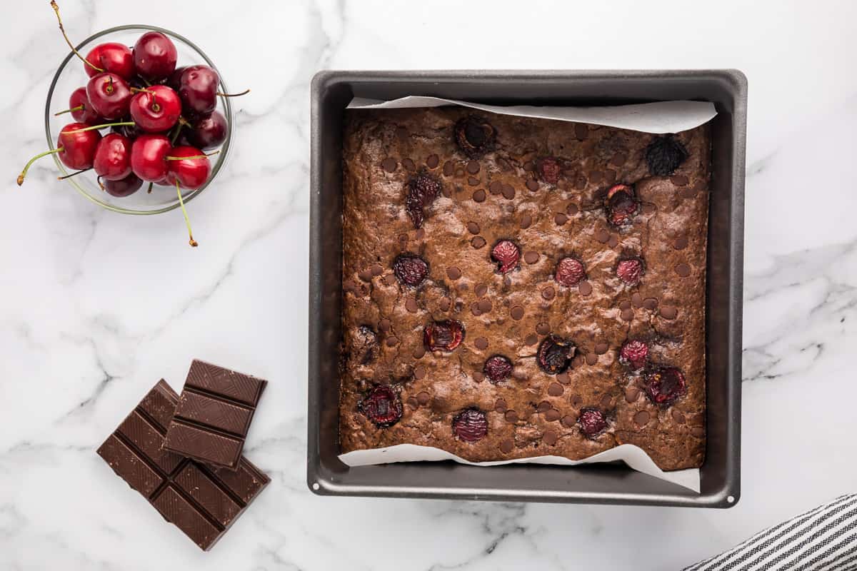 A square pan of baked double chocolate cherry brownies with cherries, chocolate and a wood spoon around it.