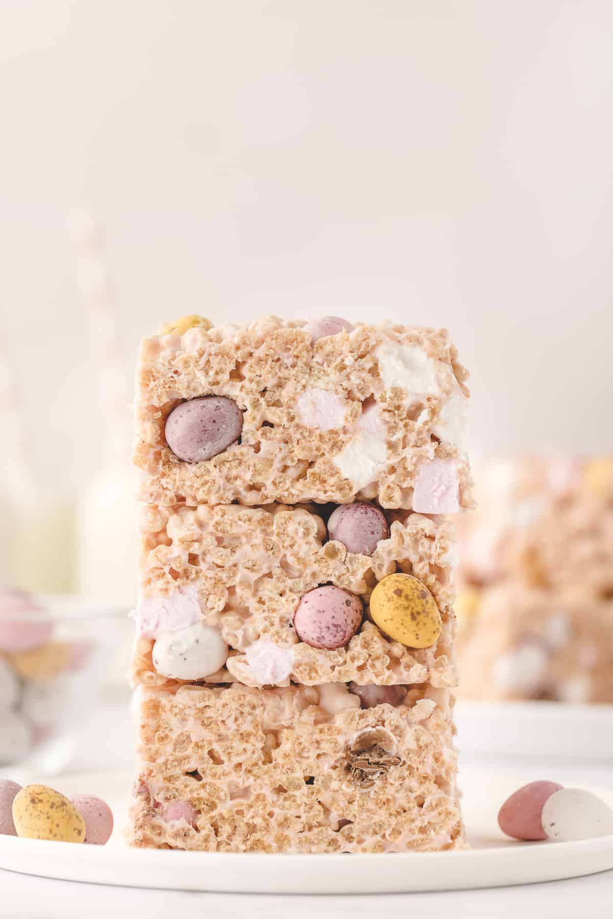 Three Easter Rice Krispies Treats stacked on top of each other with some mini candy eggs around them and more treats in the background.