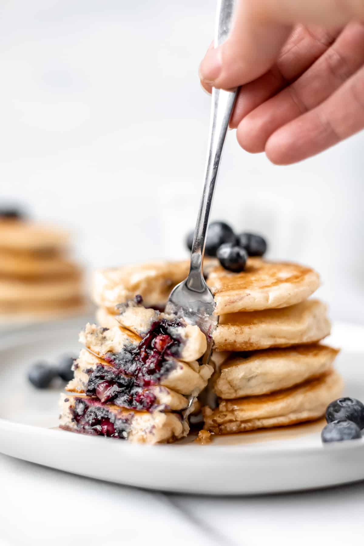 A fork with a bite of 4 stacked blueberry pancakes on it with two plates of blueberry pancakes in the background.