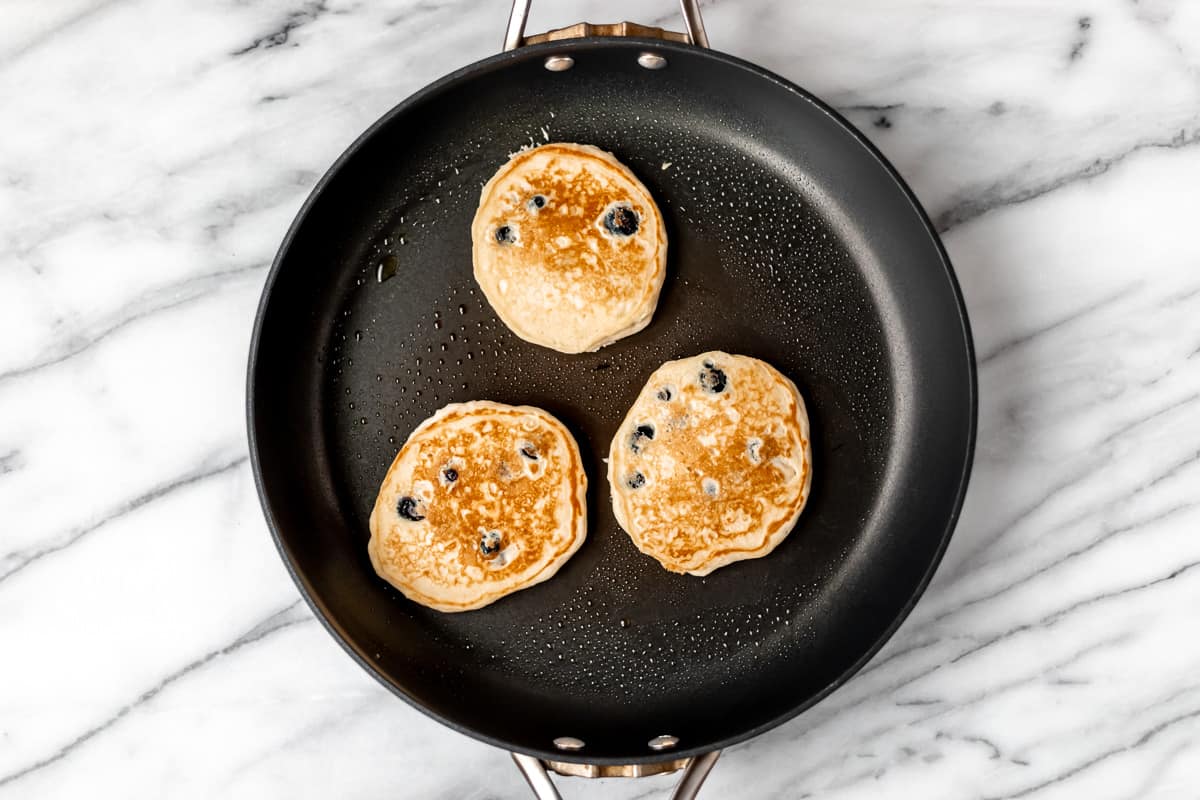 Three cooked blueberry pancakes in a black skillet over a marble background.